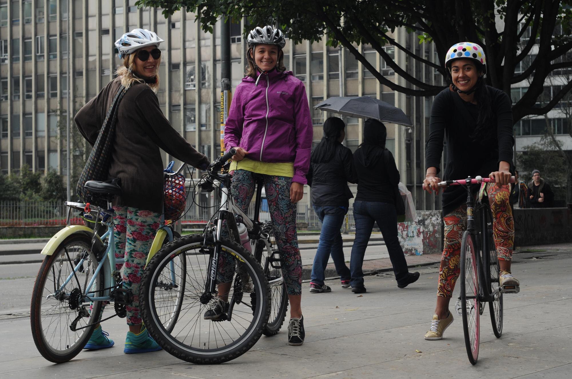 Three women cyclists wearing colorful, patterned outfits and helmets while riding through a city plaza, highlighting expressive urban cycling fashion and community culture.