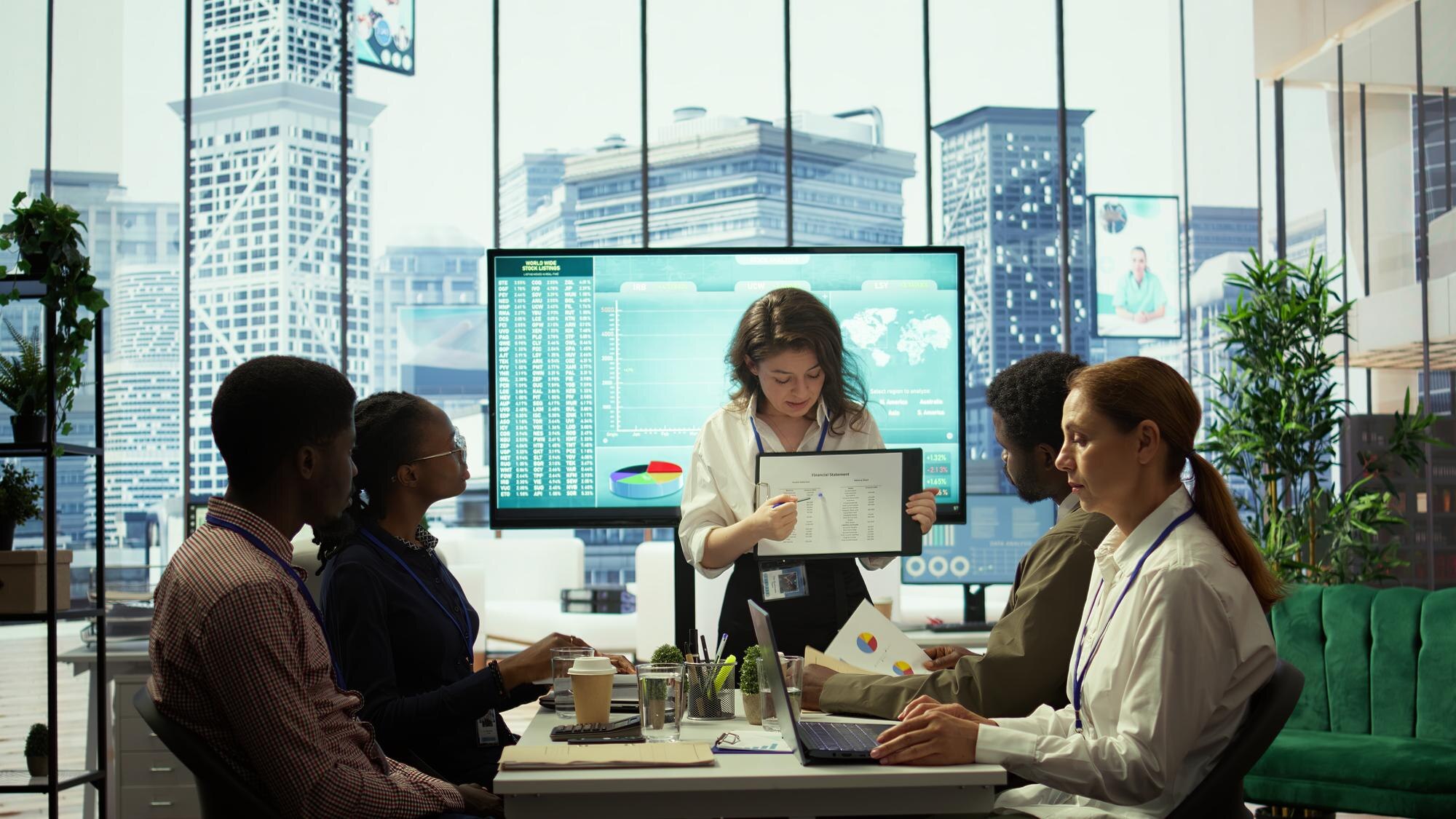 coworkers at a table watching a peer presenting data on large screen in a well lit conference room with a city view in the background