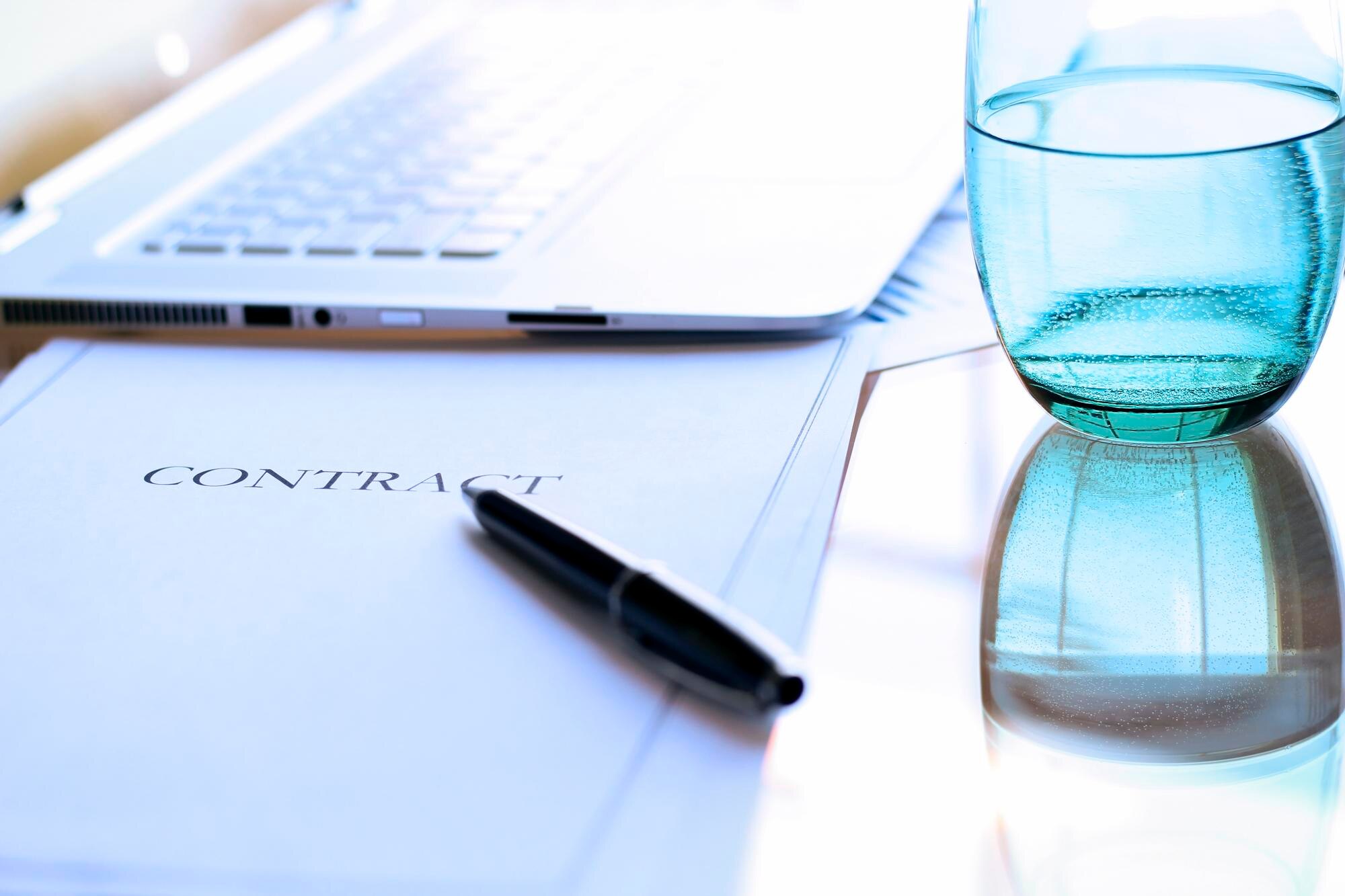 Contract document with the word "CONTRACT" visible, black pen, laptop, and a blue glass of water, illustrating business agreements and compliance essentials for startups.
