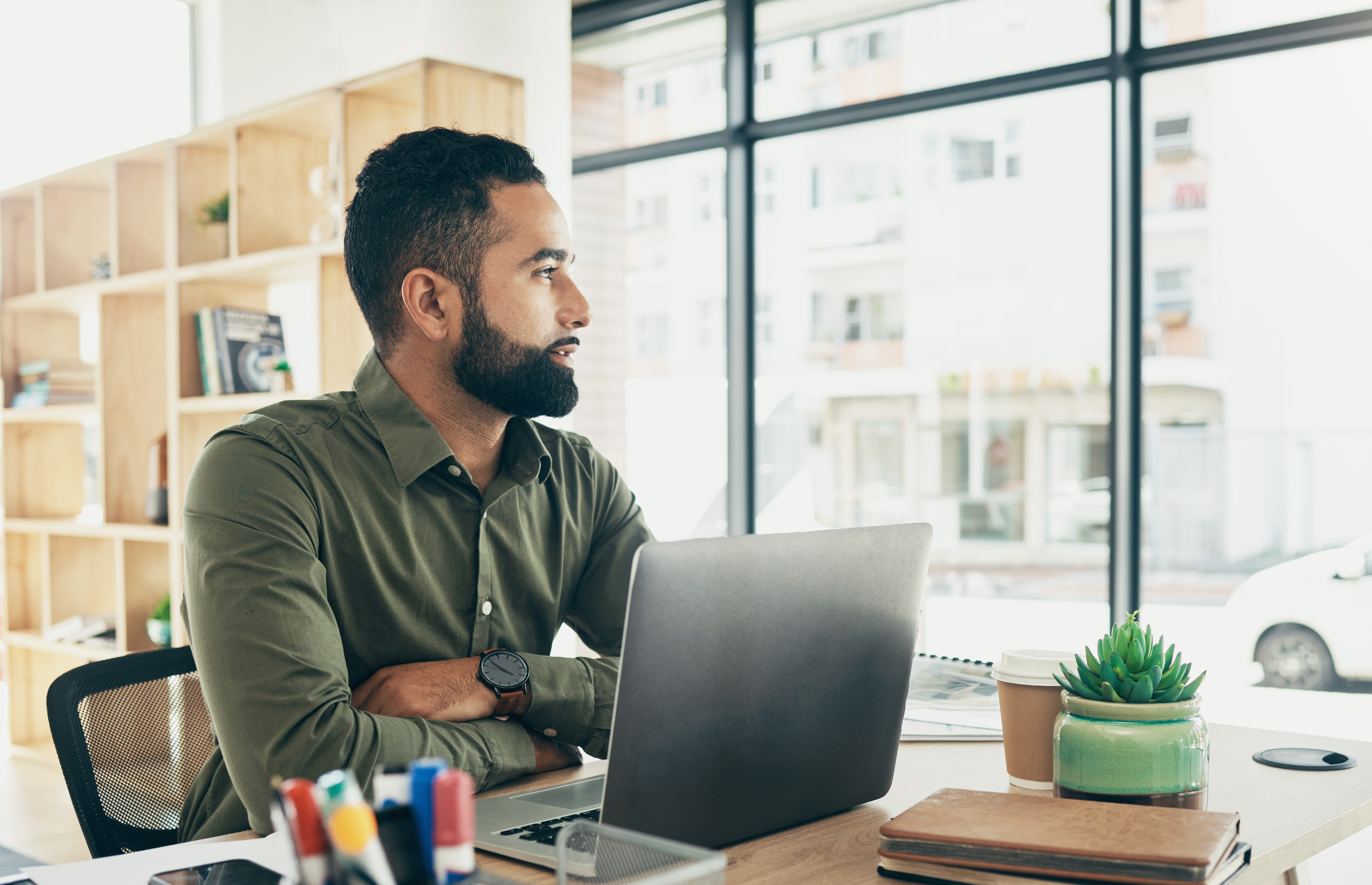 Businessman contemplating legal steps for small business success, seated at desk with laptop, stationery, and plants, in a bright office setting.