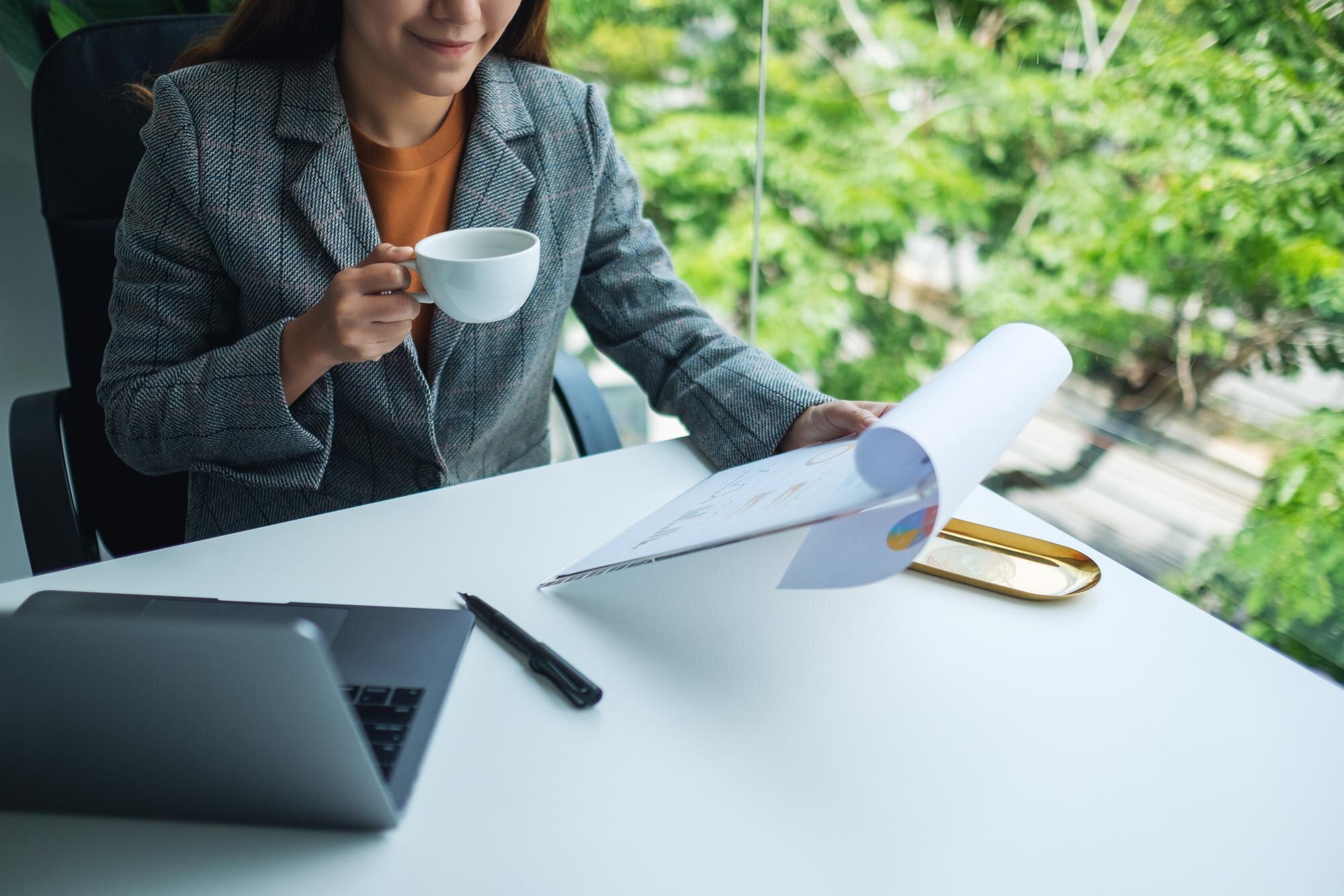 Businesswoman reviewing financial paperwork while holding a coffee cup, with a laptop and a green outdoor view in a modern office setting.
