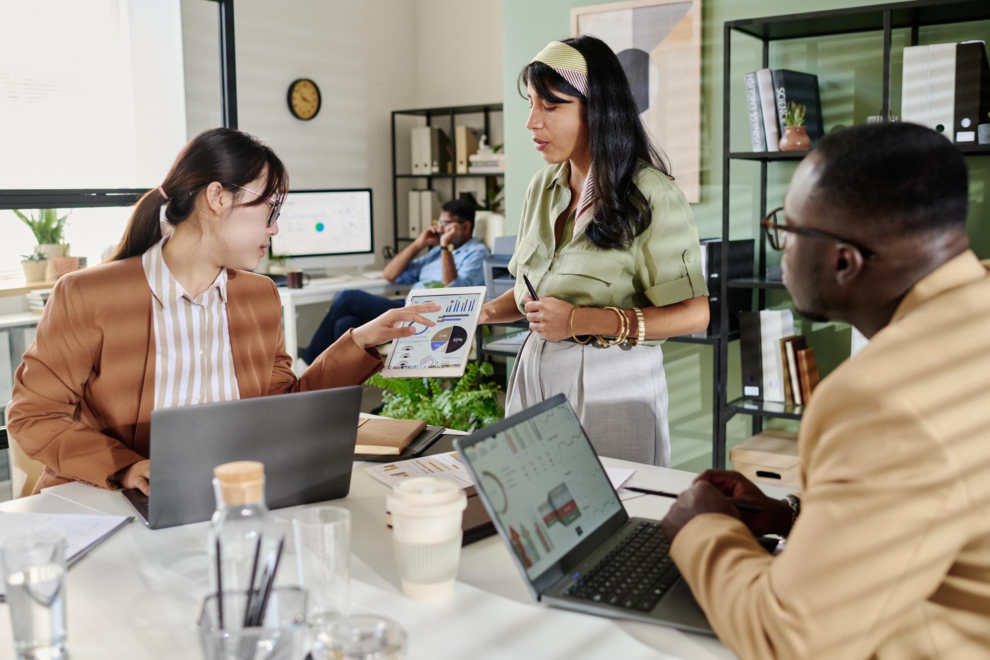 Team members collaborating in a modern office, discussing data insights on a tablet, with laptops and business documents on the table, illustrating effective communication and teamwork for SEO strategy enhancement.