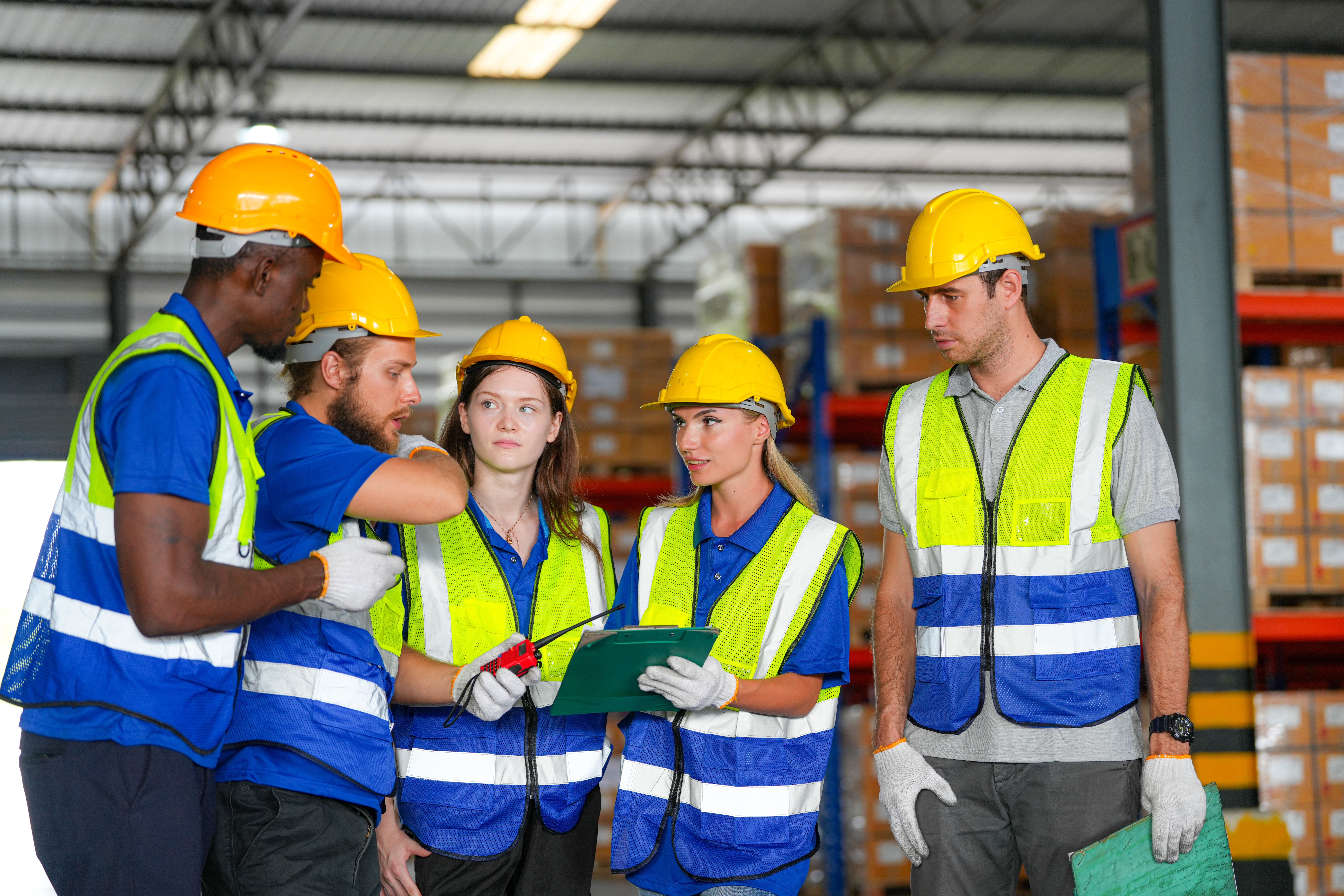 Een groep medewerkers in een magazijn bespreekt samen een werkinstructie, uitgerust met veiligheidshesjes en helmen.