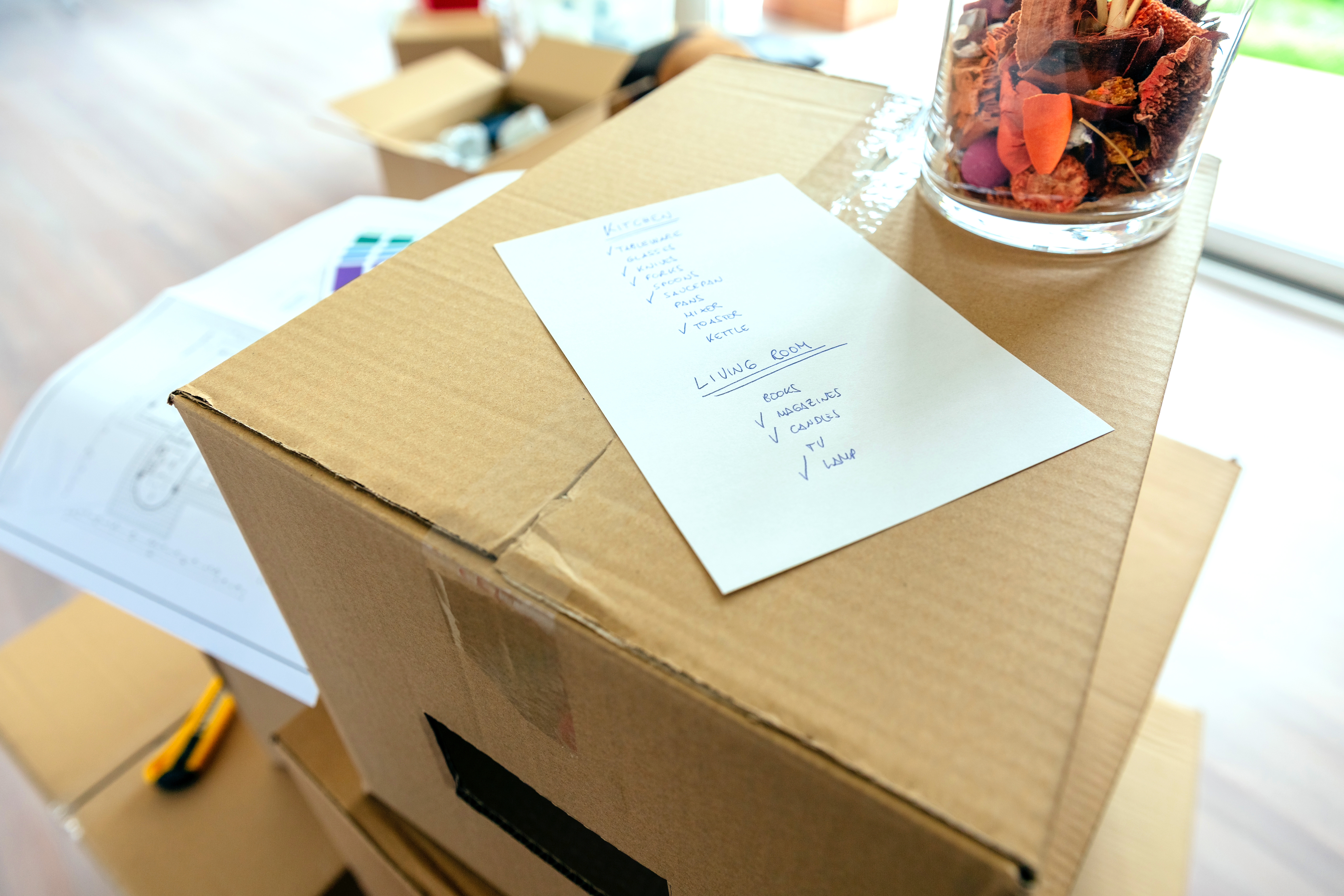 A packing checklist, listing rooms and items within each room, lays on top of a stack of boxes. Next to the list, also on top of the box, is a jar of potpourri.