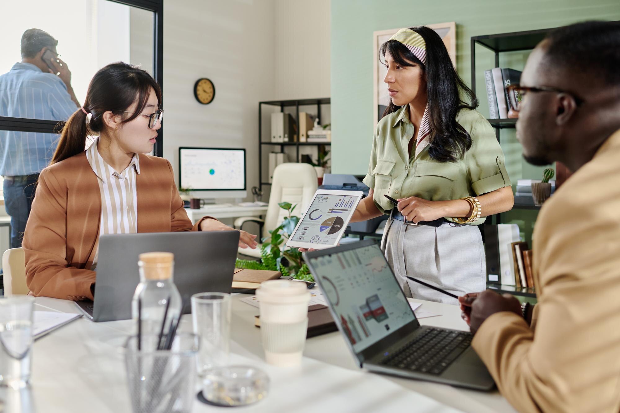 Business meeting in modern office, discussing strategies with laptops and tablet displaying graphs, focus on collaboration and productivity.