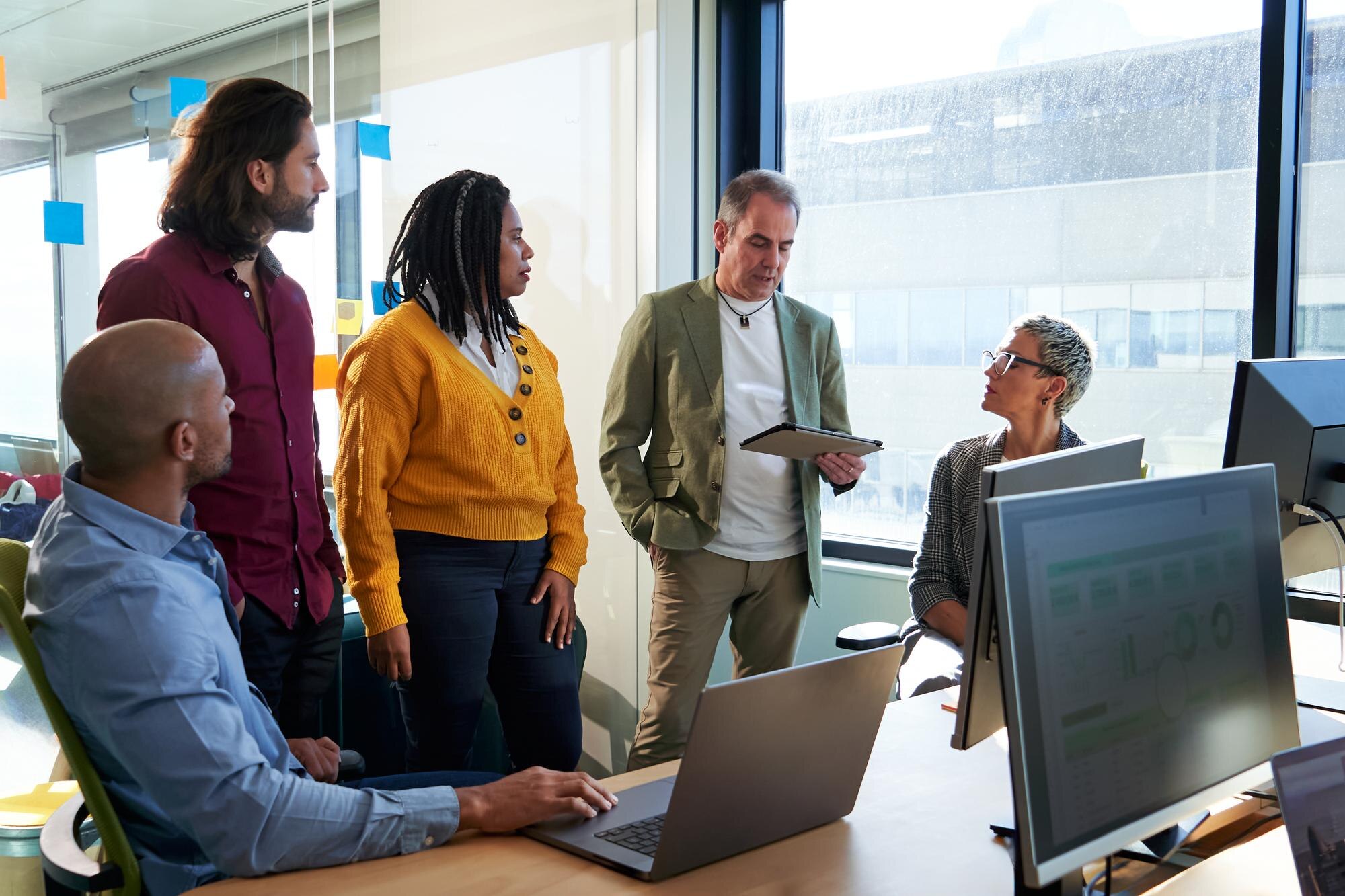 Diverse business team collaborating in a meeting, discussing local SEO strategies and consistency in business information, with laptops and monitors displaying data.