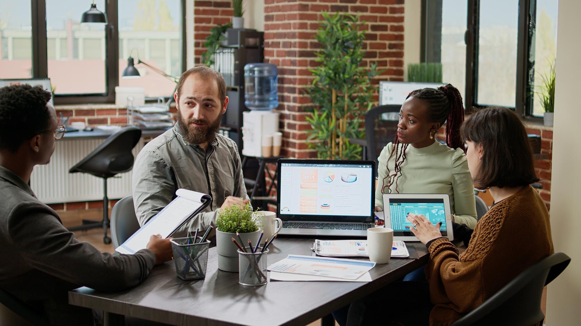 Diverse team of business professionals collaborating on research and strategy, discussing insights with laptops and tablets in a modern workspace.