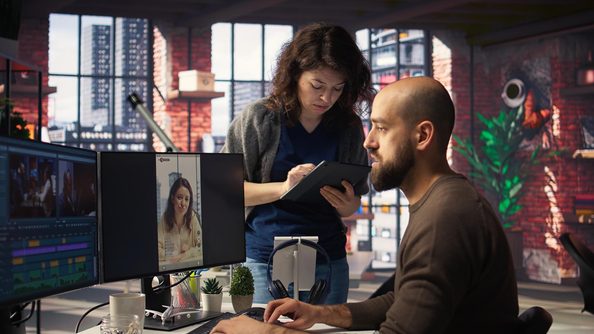 Two professionals collaborating in a modern workspace, one editing video content on dual monitors while the other takes notes on a tablet, highlighting the use of videography software for business engagement.