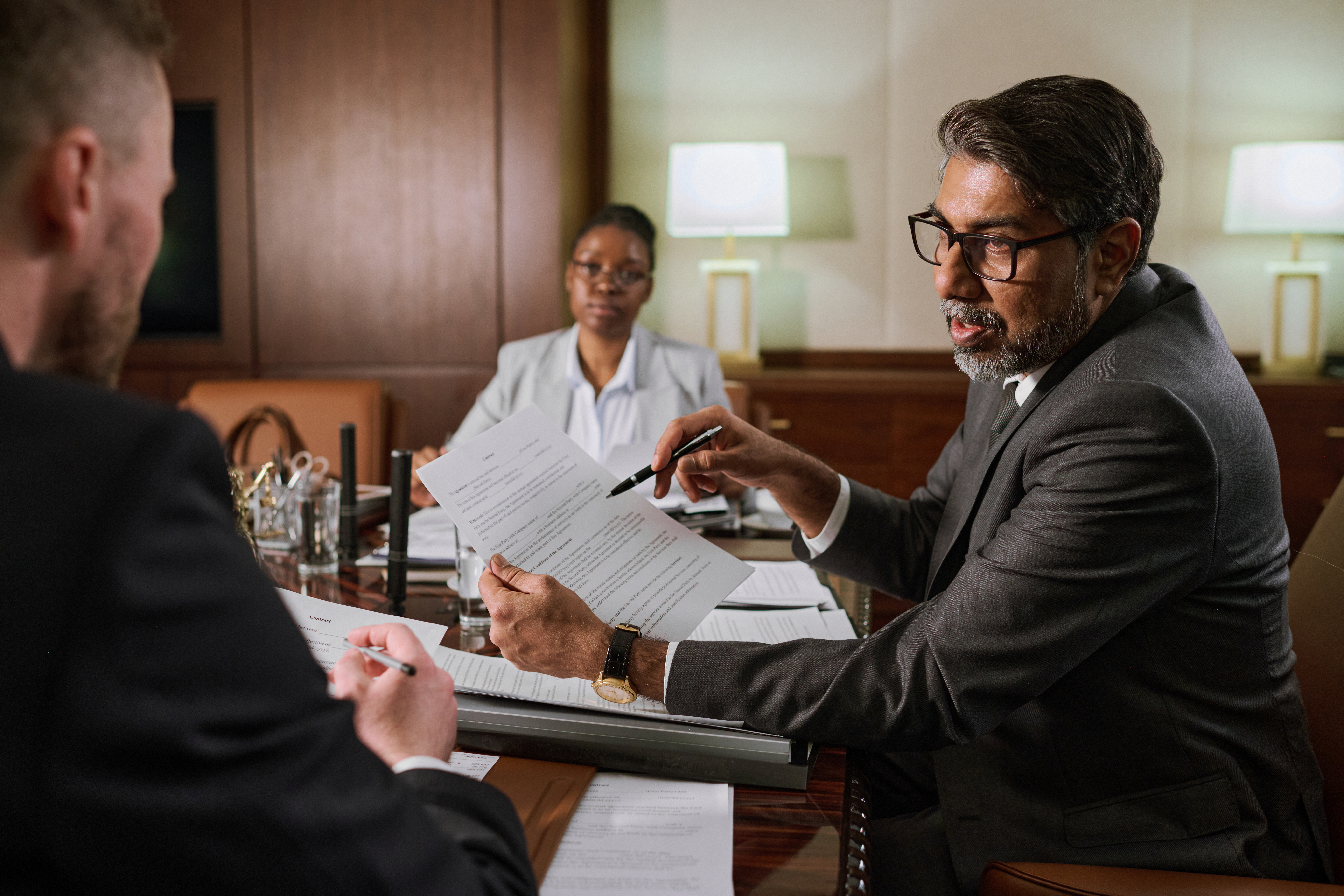 Business meeting discussing estate planning documents, with a man in a suit pointing at paperwork, a woman in a white blouse listening attentively, and another person partially visible, emphasizing the importance of legal guidance in asset protection.