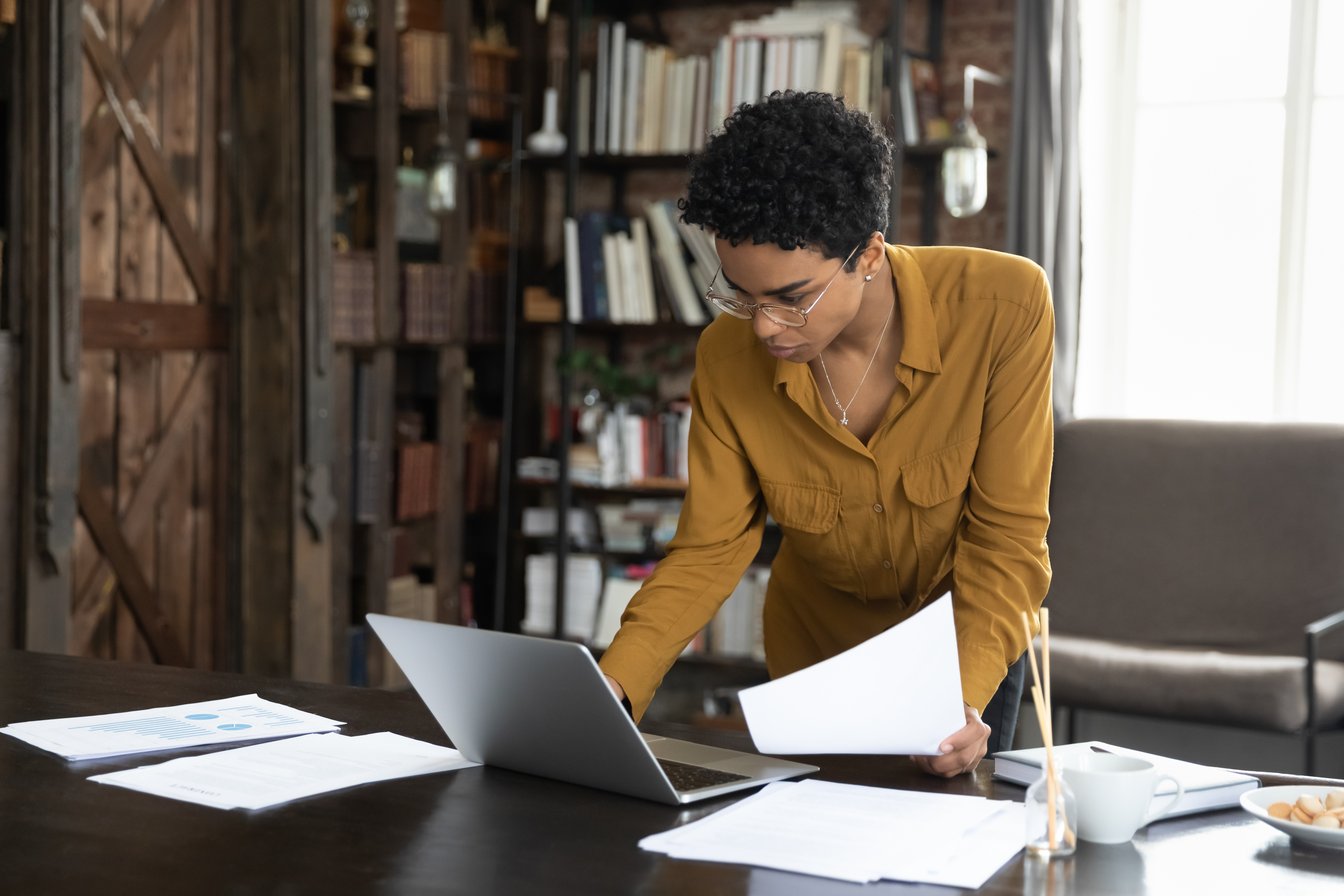 Focused young Black businesswoman reviewing documents and working on a laptop in a home office setting, emphasizing legal compliance and business management.
