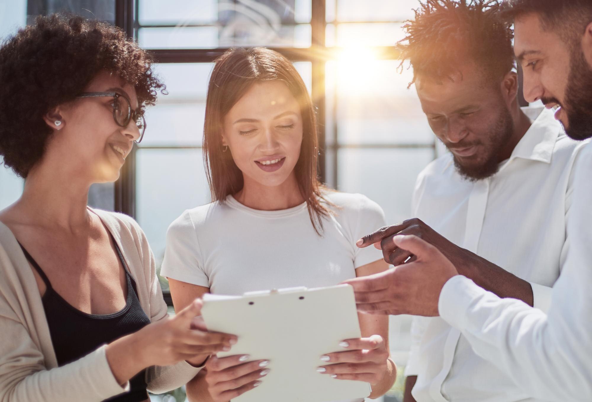 Young diverse team discussing project details on a tablet, smiling and engaging in collaborative conversation, in a bright office setting, emphasizing teamwork and customer feedback.