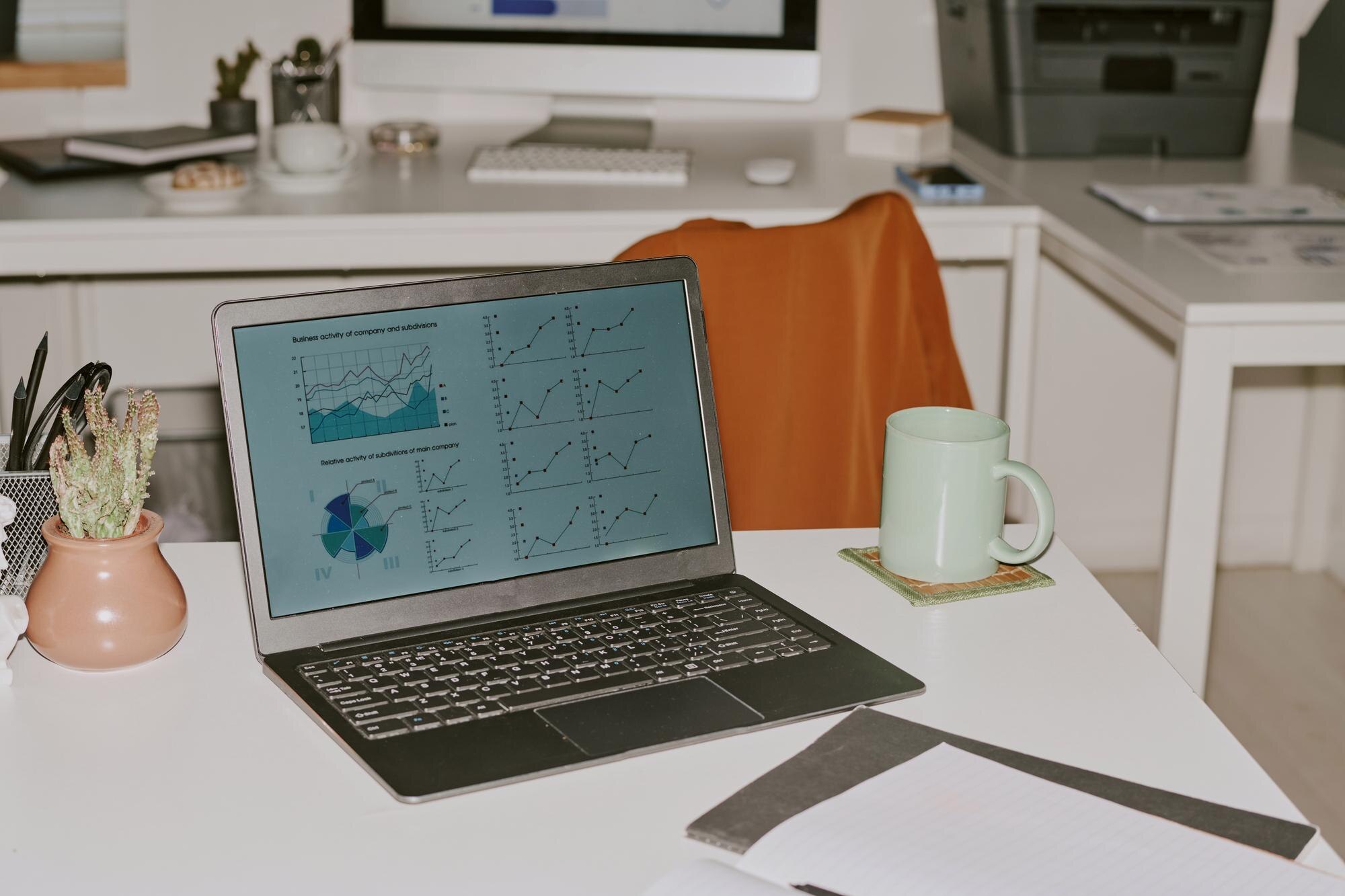 Laptop displaying business activity graphs and charts in a modern workspace, with a green mug and notebook nearby, emphasizing effective digital marketing strategies for improved user engagement.