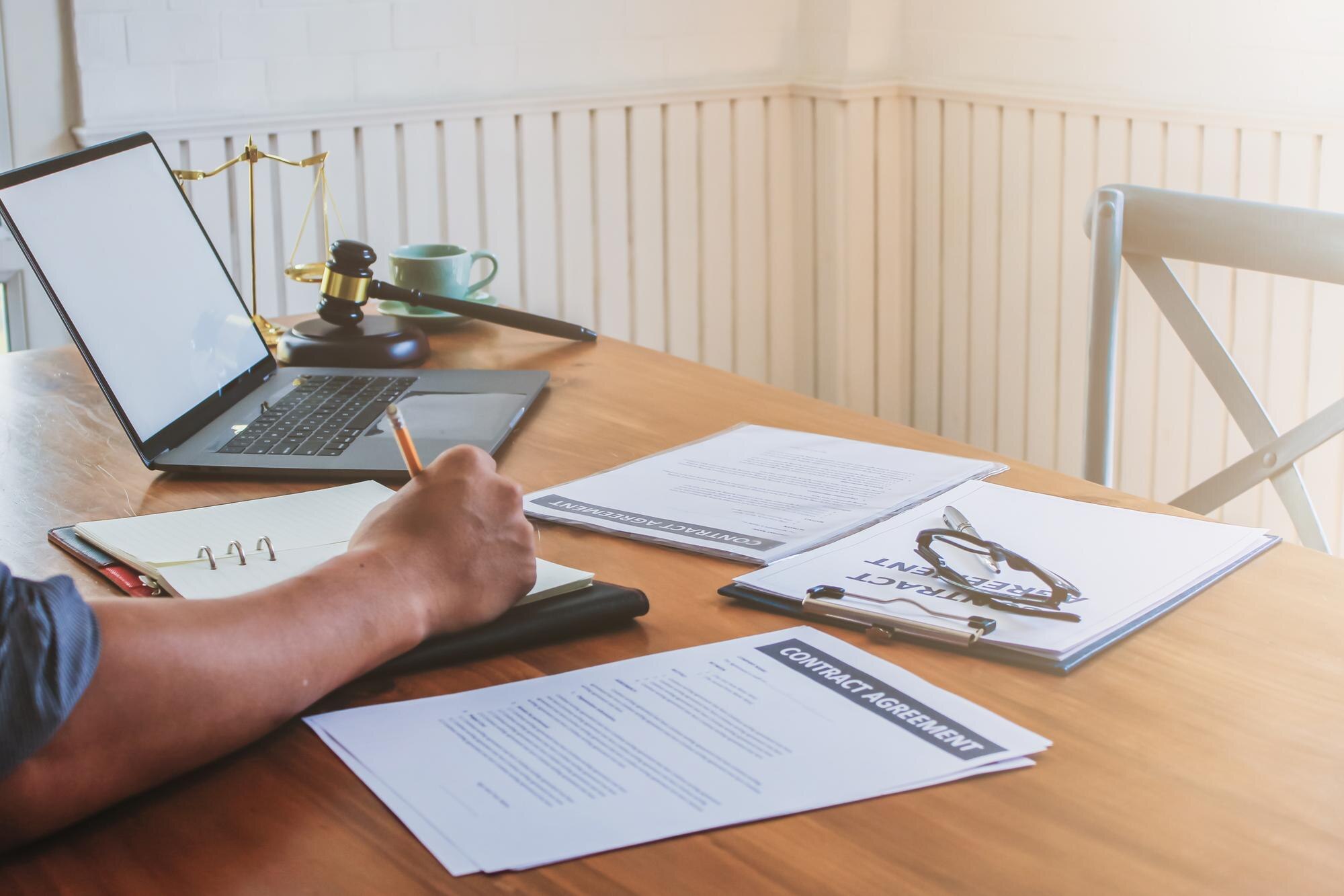 Person writing in notebook at a desk with laptop, legal gavel, and contract agreements, illustrating essential legal documentation for startups.