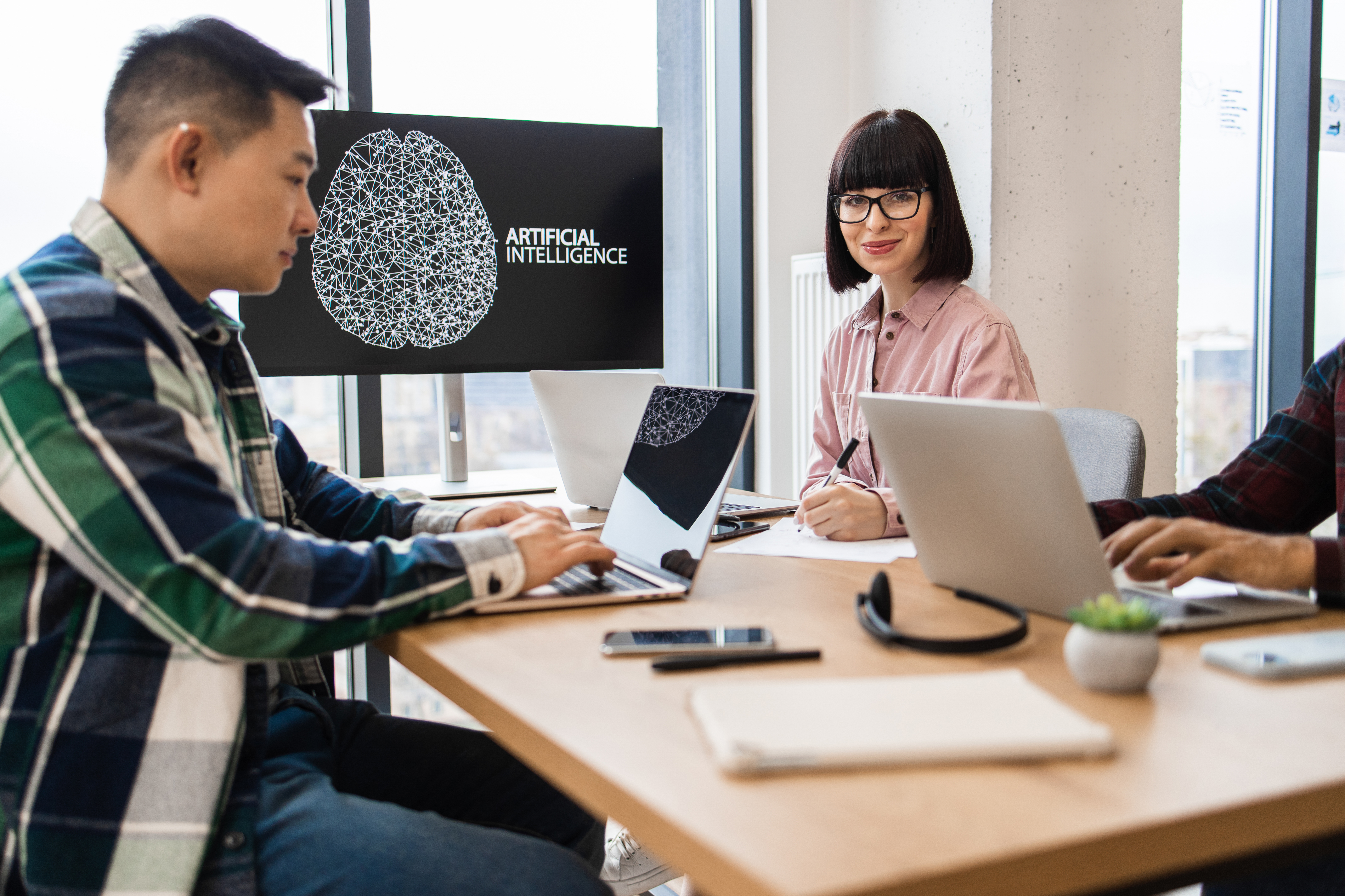 Team of IT experts collaborating on artificial intelligence development, with a monitor displaying "ARTIFICIAL INTELLIGENCE" and laptops on a table, highlighting the integration of AI in business solutions.