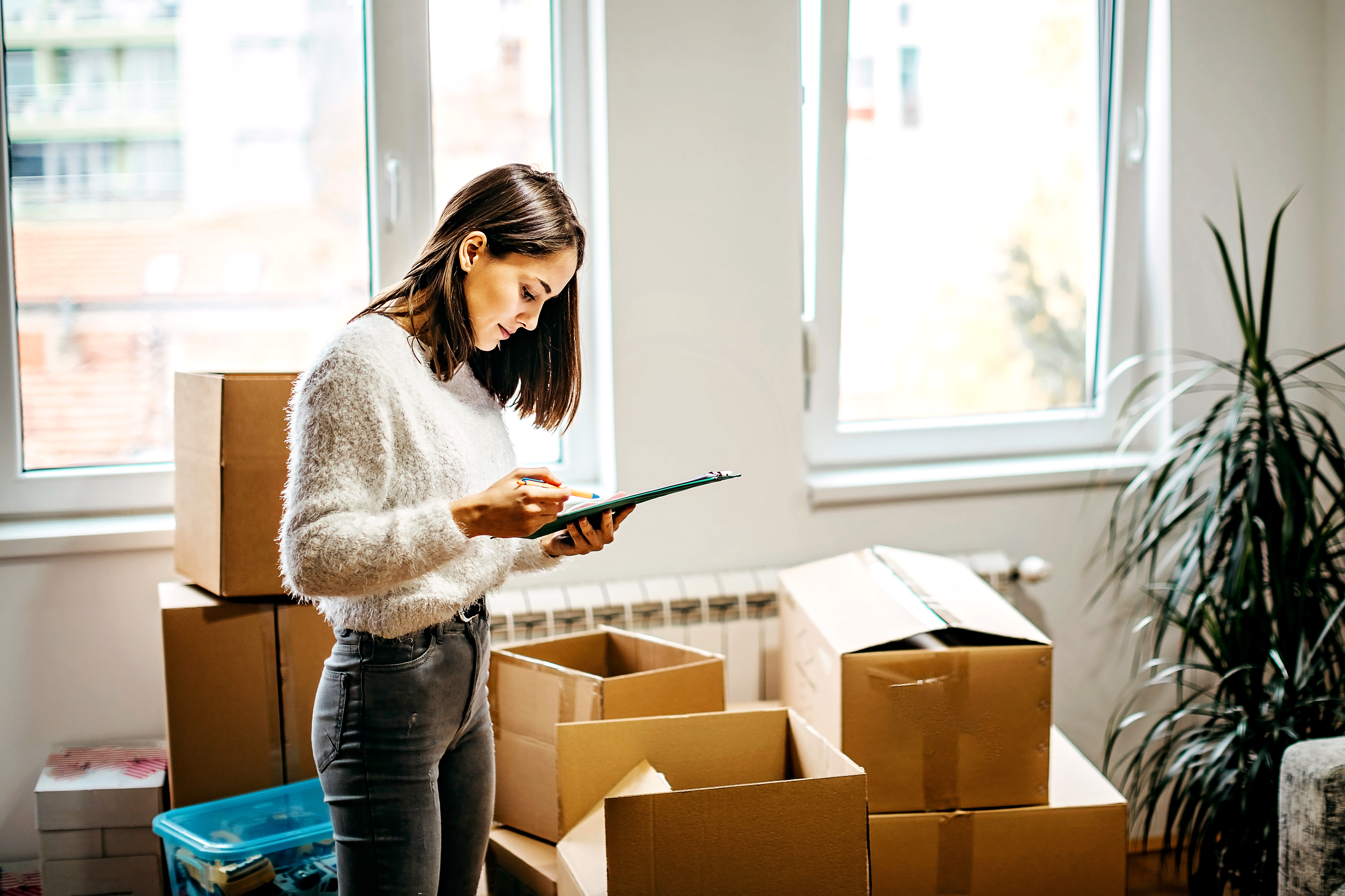 A woman stands in a room amongst empty boxes. She looks down at a clipboard, the clipboard in one hand and a pencil in the other.
