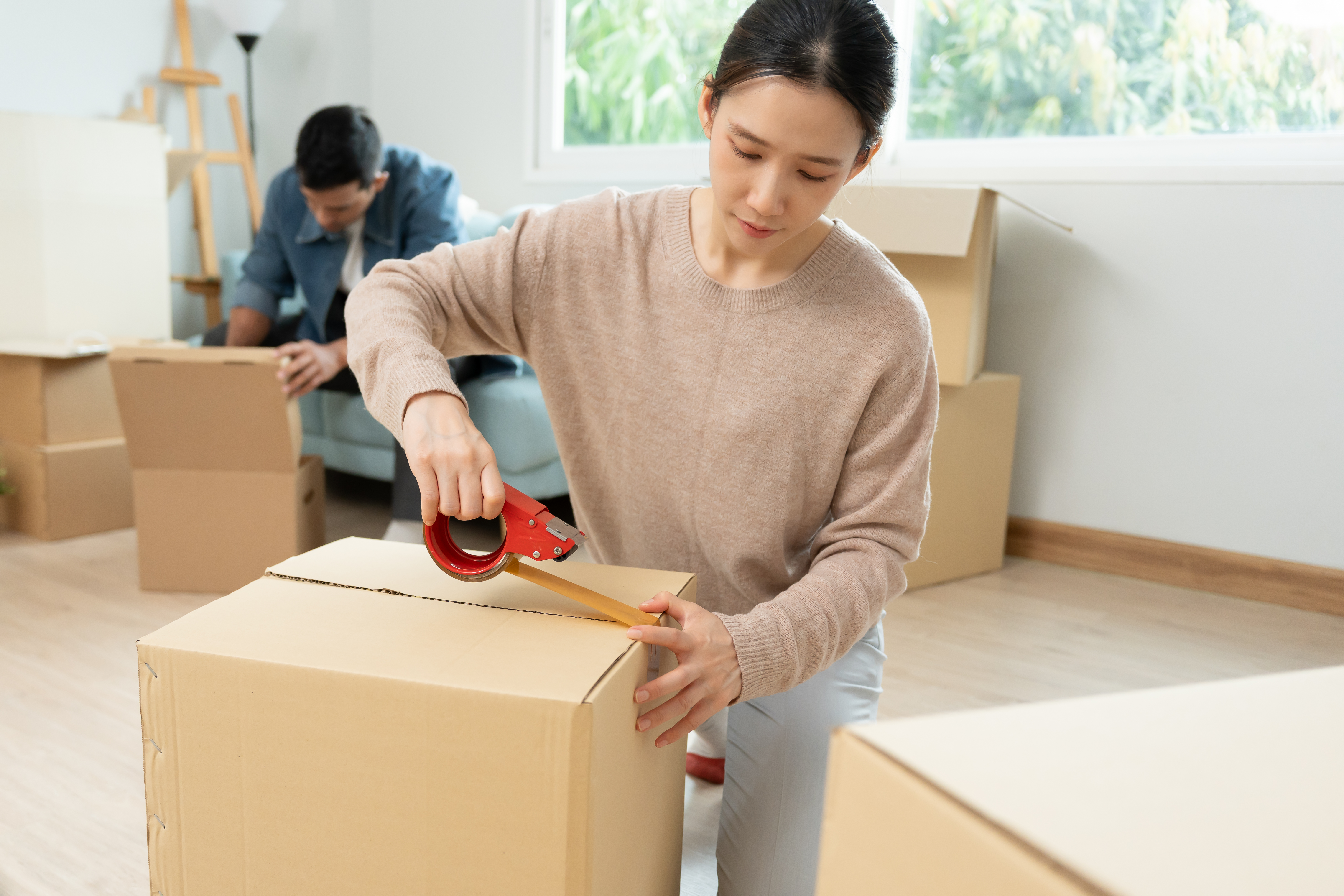A woman kneels on the floor, packing a box shut with packing tape. A man sits further behind her on a couch, looking inside a box. There are other open and closed boxes stacked around them.