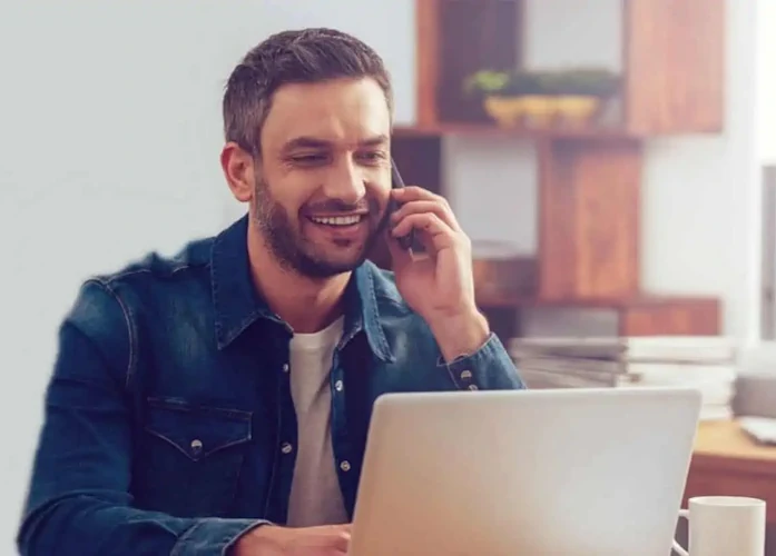 Man smiling while talking on phone, laptop and coffee nearby.