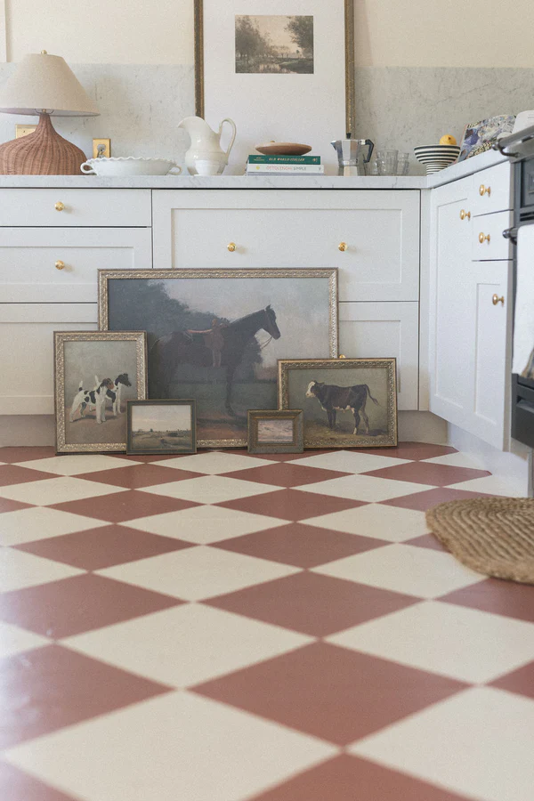 An assortment of vintage inspired artwork featuring farm animals and landscapes resting against a cupboards in a orange and white checkered floor in a farmhouse kitchen.