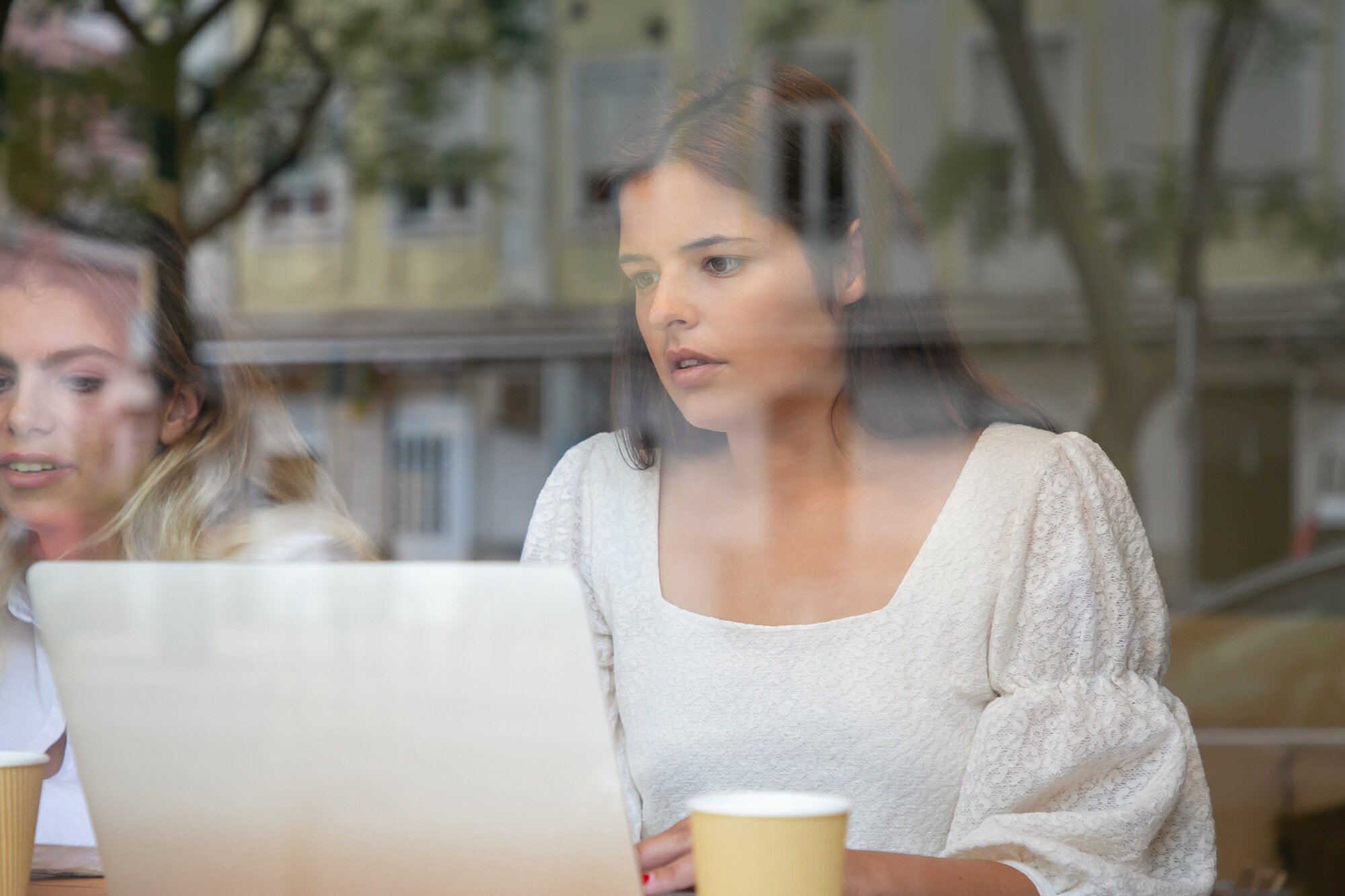 Brunette woman focused on laptop, discussing Google Business Profile management with a blonde woman, coffee cups on table, in a café setting.