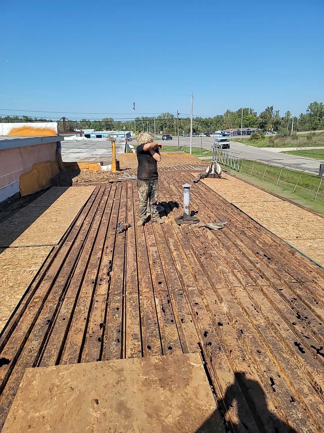 an old roof torn down to the decking with the old decking removed and new decking being installed in areas of the picture. A person wiping sweat from their brow is in the picture.