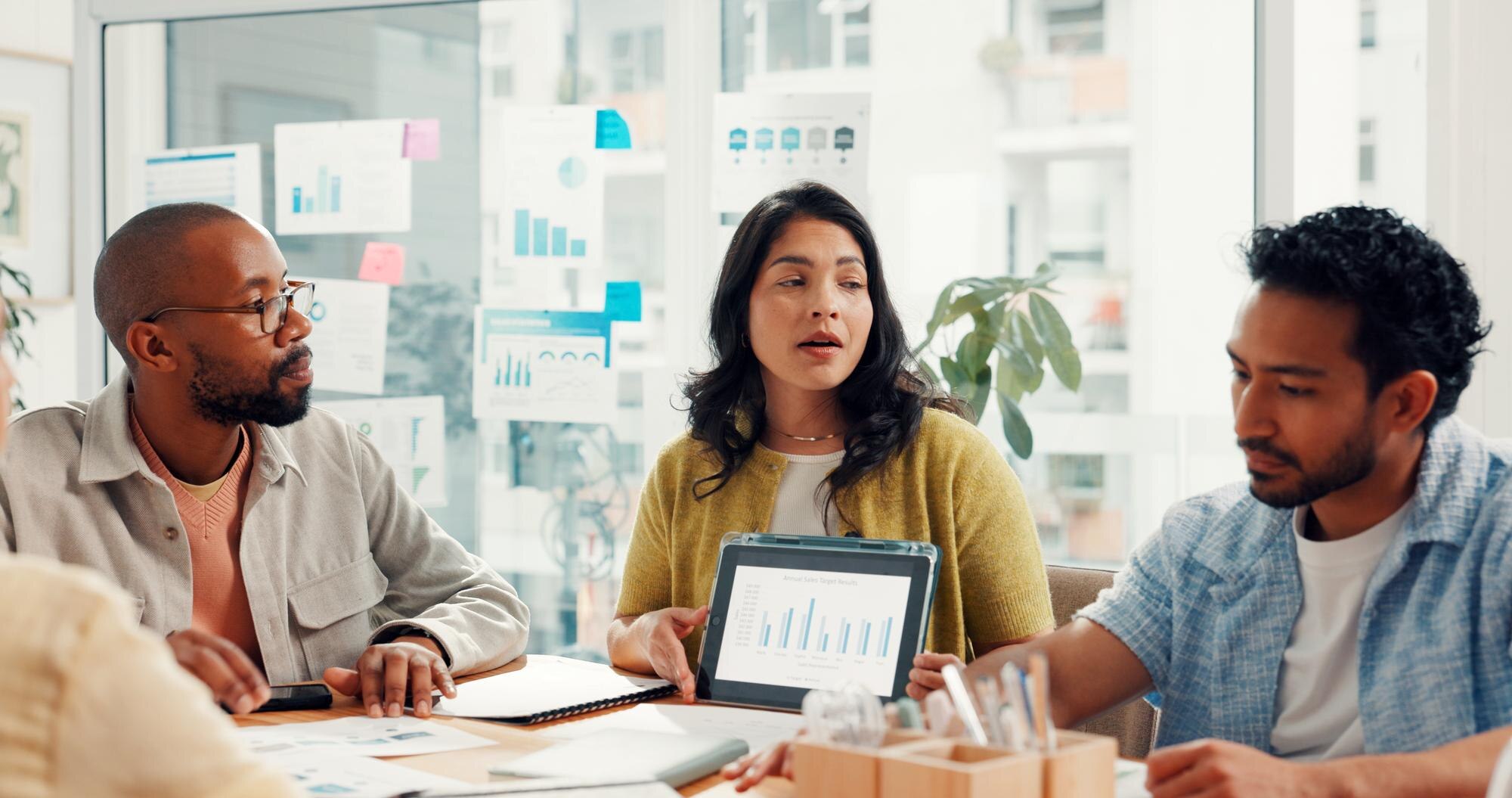 Business meeting with diverse professionals discussing research data on tablet, surrounded by charts and graphs, emphasising the importance of accurate business information for local SEO success.