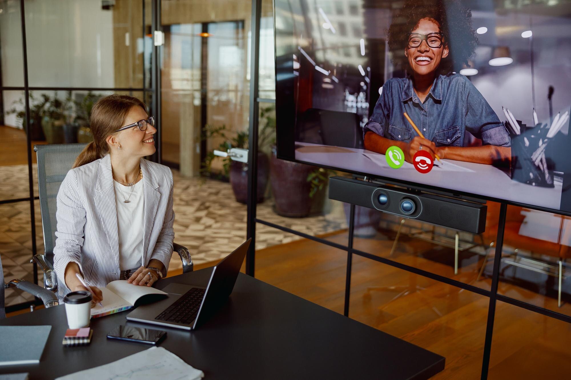 Professional woman in a contemporary office engaged in a video conference with a smiling participant on a large screen, illustrating the power of video communication for business interactions.