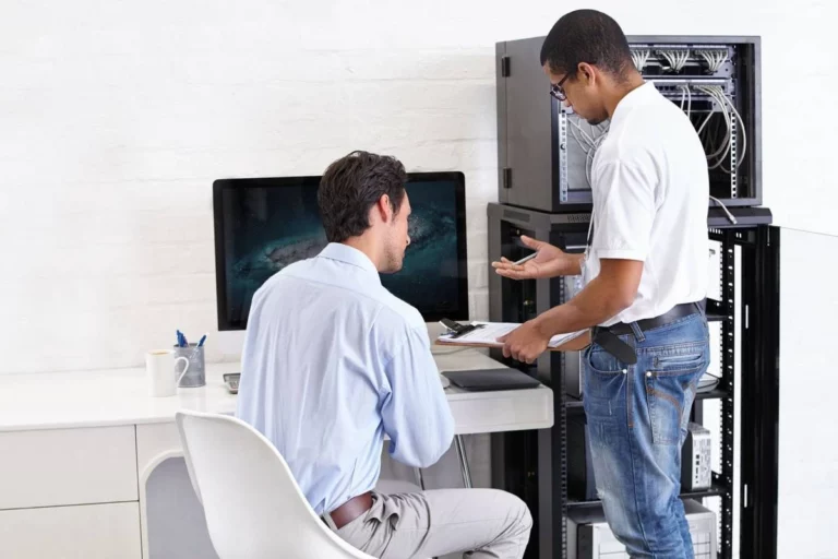 Two professionals discussing IT support strategies near a computer and server rack, emphasizing business continuity and disaster recovery planning.